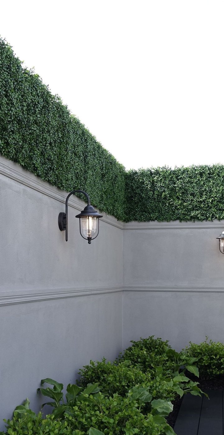 Narrow courtyard with rendered grey walls topped by a dense green hedge and lit by lantern wall lights