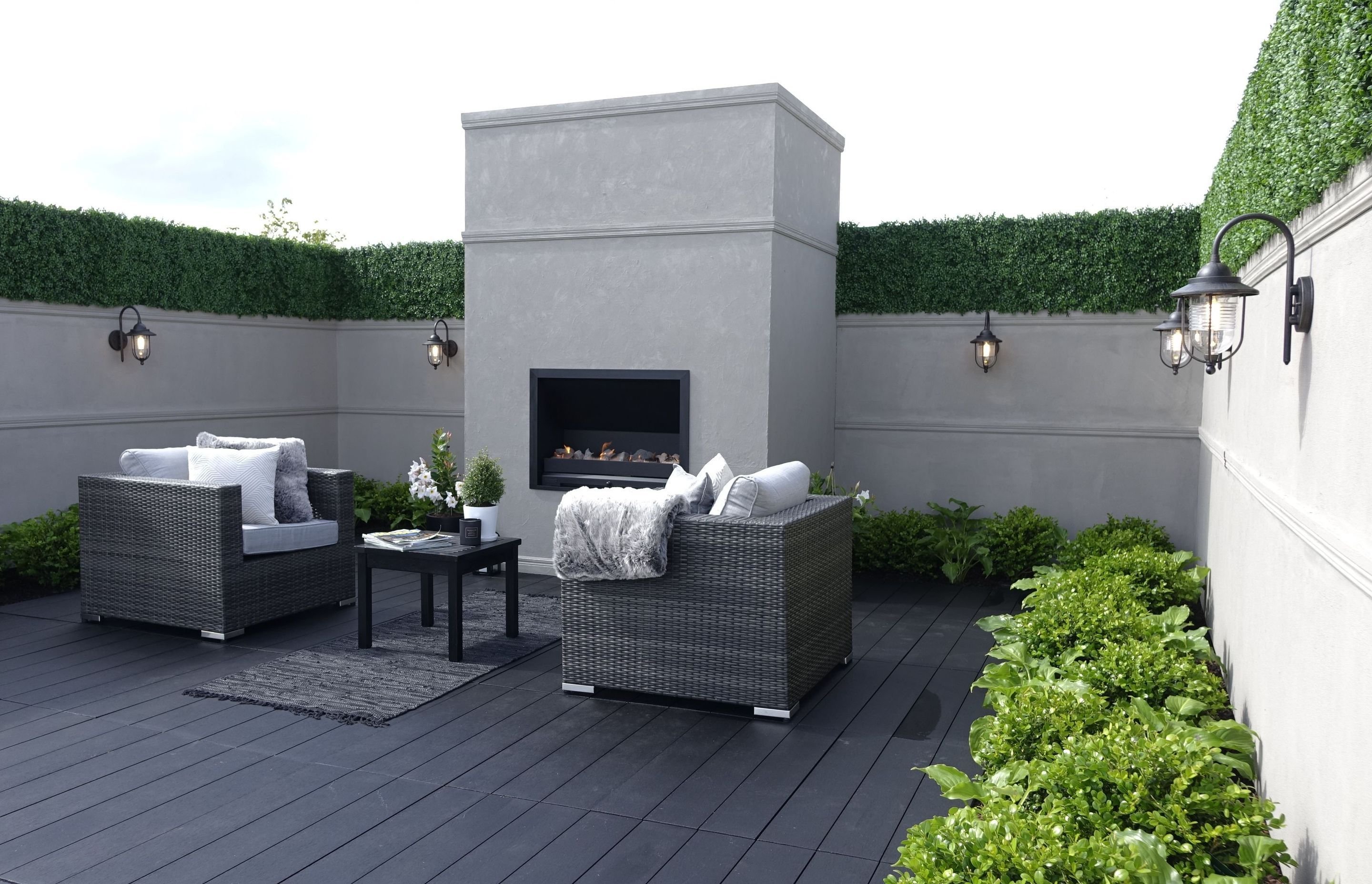 Grey courtyard with a rendered outdoor fireplace, wicker lounge seating and a dense green hedge along the wall