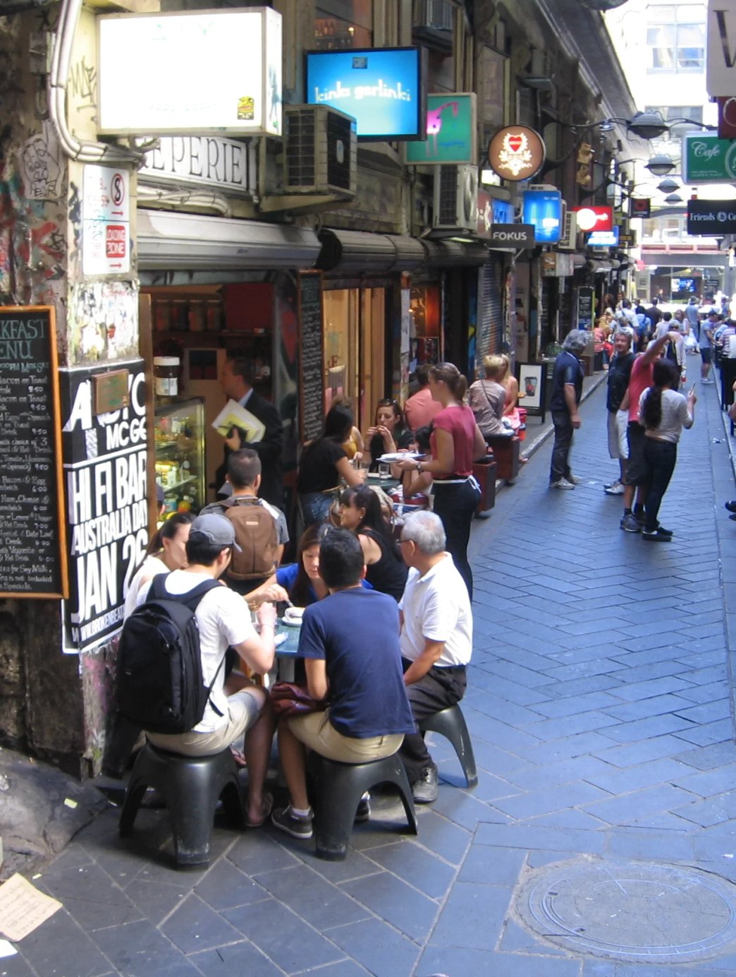 A busy Melbourne laneway lined with cafe tables, street signage and a busker playing accordion