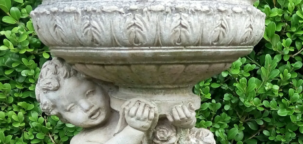 A classical stone urn planter carried by a cherub, set against a clipped buxus hedge