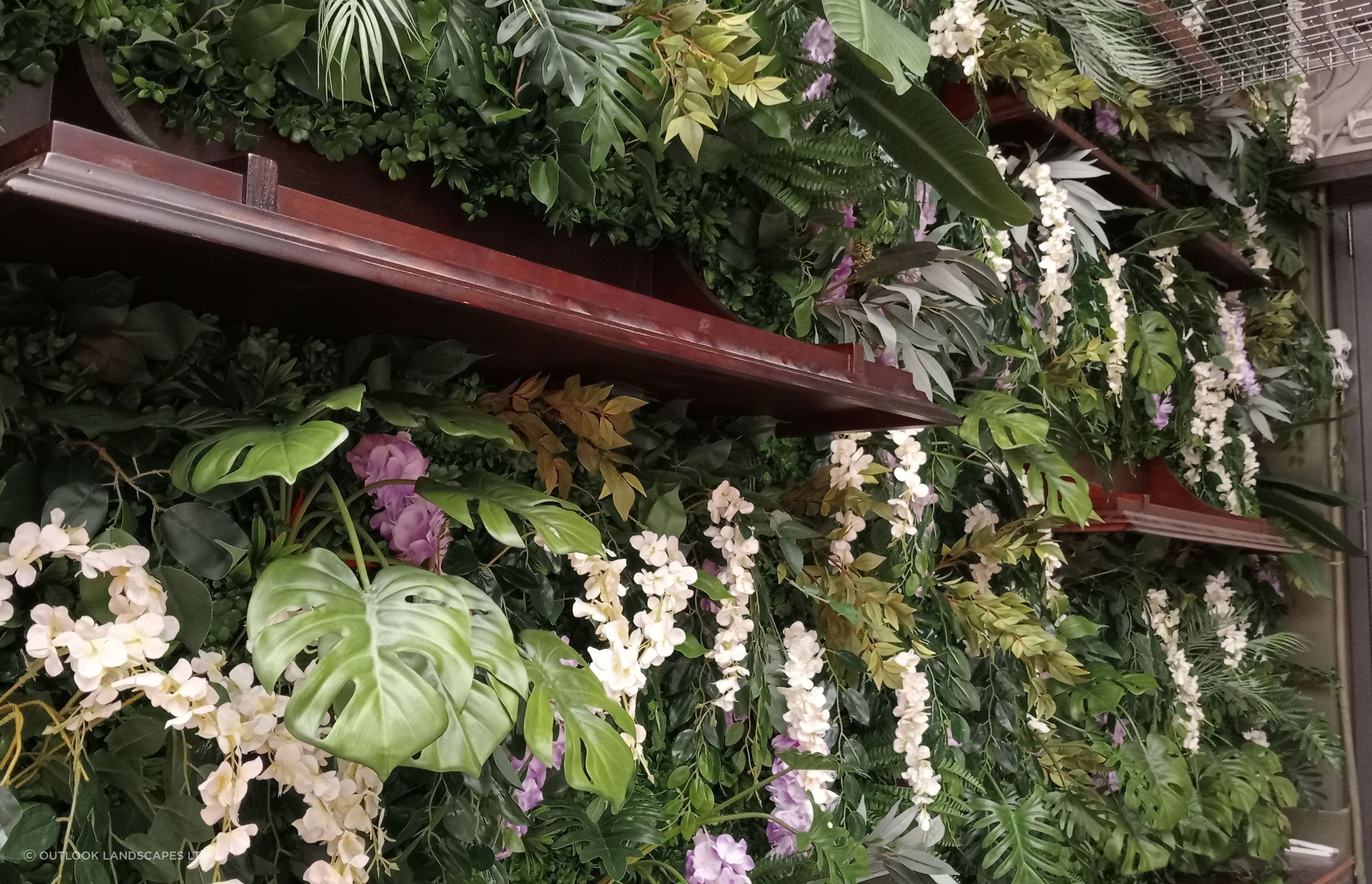 Artificial green wall of mixed foliage and trailing white and lilac blooms, framed by dark timber shelving
