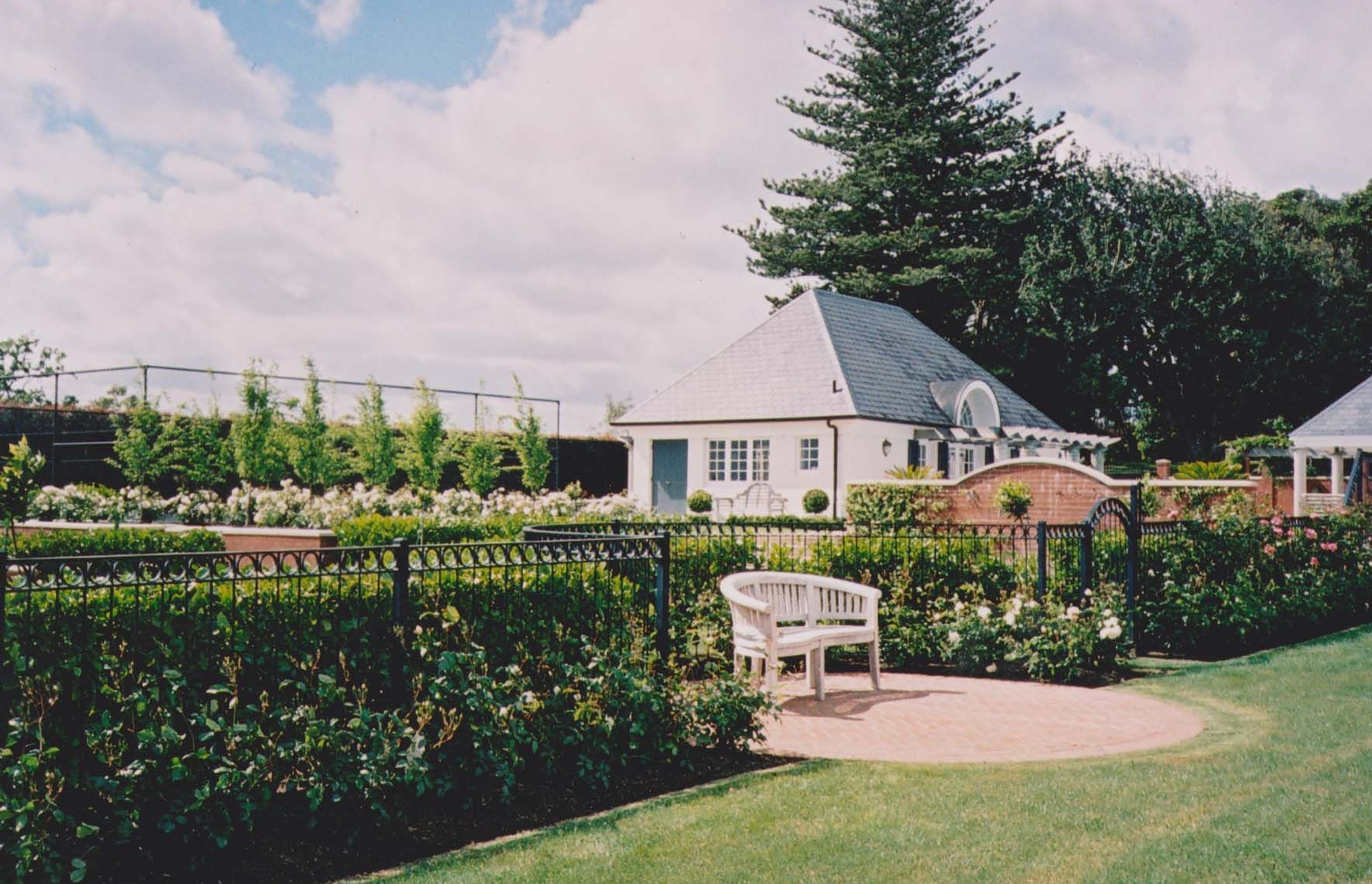 Formal rose garden with clipped hedging, a white timber bench on brick paving and a white pavilion beyond